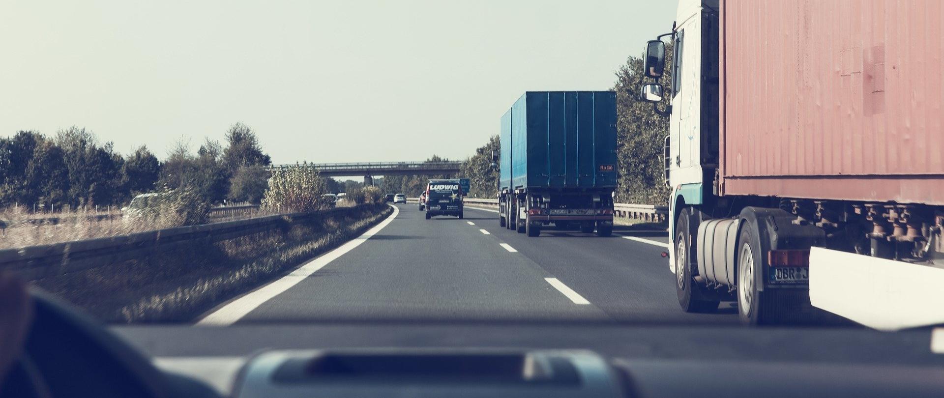 Zdjęcie przedstawia samochody jadące po drodze szybkiego ruchu. Na prawym pasie jadą dwa ciężarowe pojazdy. Na lewym, na dalszym planie, samochód osobowy. Zdjęcie wykonane z perspektywy osoby prowadzącej samochód.