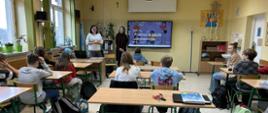 Dwie kobiety stoją obok tablicy multimedialnej w zali szkolnej. W ławkach siedzą dzieci