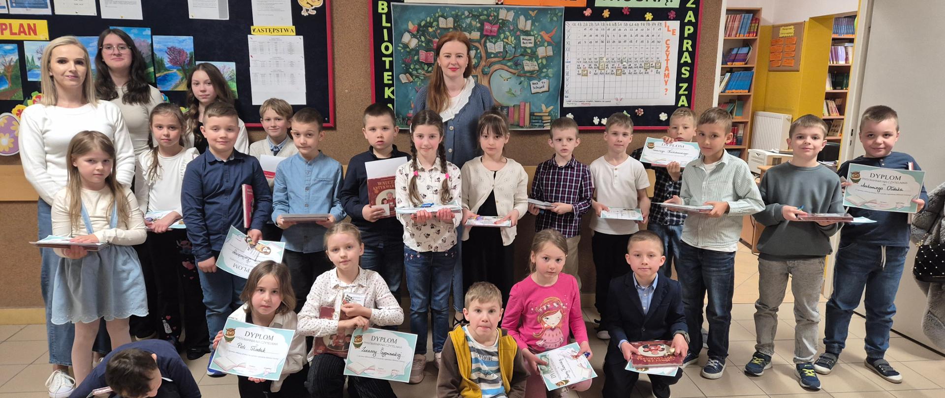 Grupa dzieci. Stoją a ci z przodu kucają. Trzymają w rękach dyplomy oraz czerwone książki. Za nimi stoi kobieta a dwie inne z boku. Na ścianie za nimi napis Czytanie kwitnie wiosną