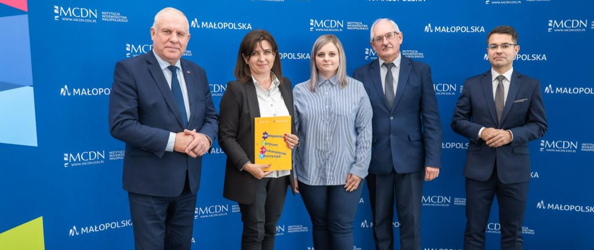 Na granatowym tle ścianki do zdjęć stoją elegancko ubrani mężczyźni i kobiety. Jedna z kobiet trzyma w ręku teczkę z podpisaną umową.
