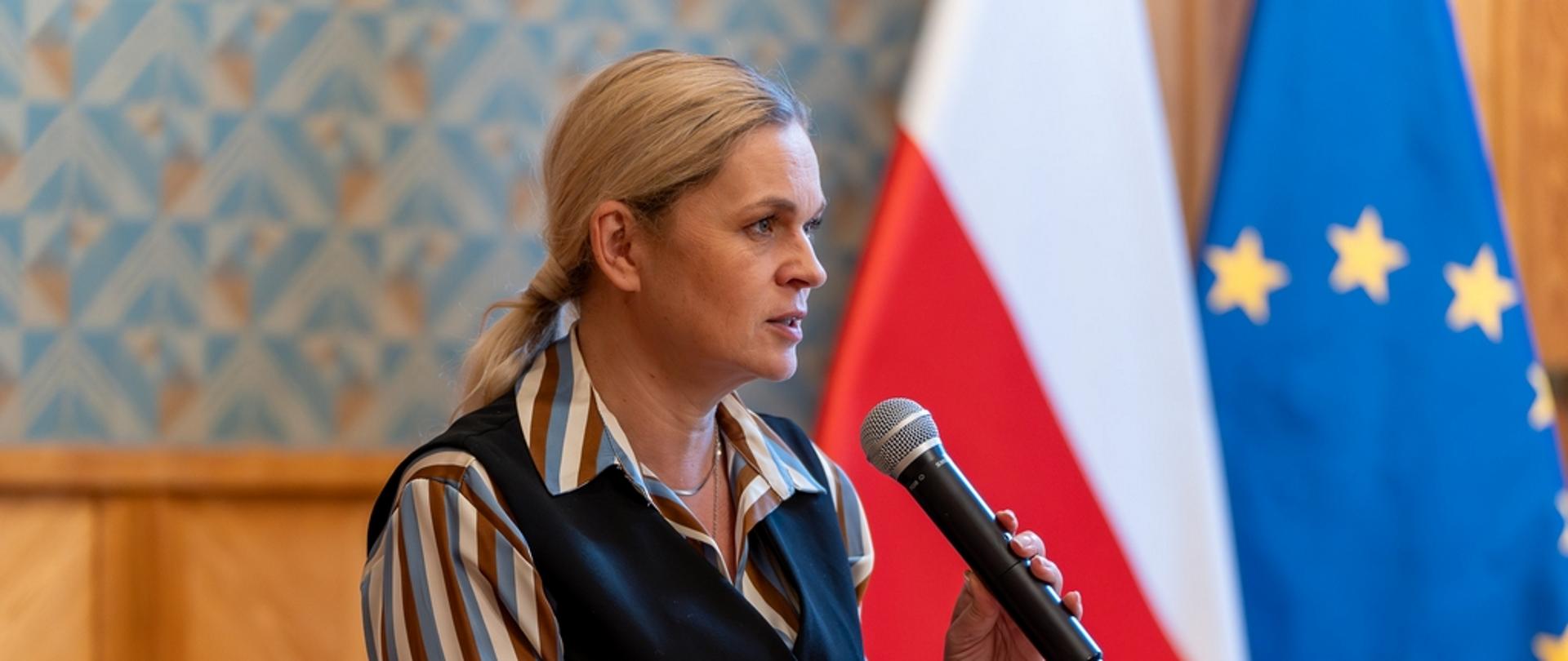 Ministra Edukacji Narodowej Barbara Nowacka trzyma w ręku mikrofon i przemawia. W tle flaga Polski i Unii Europejskiej.