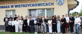 Liczna grupa dzieci, młodzieży i dorosłych pozuje przed budynkiem biblioteki publicznej; część uczestników trzyma książki i materiały z wydarzenia.