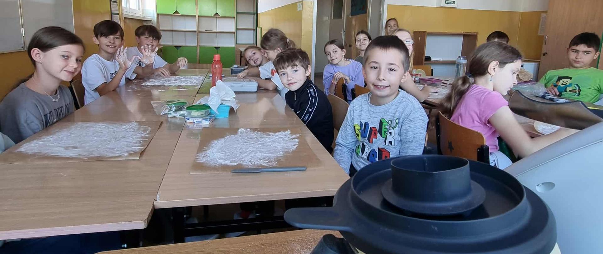 Dzieci siedziące wspólnie przy dwóch stołach, czekające na wyrabianie ciasta na bułeczki. Na pierwszym planie w misce wyrośnięte ciasto drożdżowe.