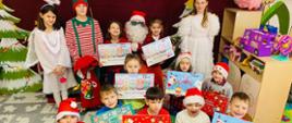 Uczniowie klasy pierwszej pozują do zdjęcia z mikołajkowymi prezentami, z tyłu siedzi Mikołaj, trzyma dwie dziewczynki na kolanach, po bokach stoją Aniołki i Elf
