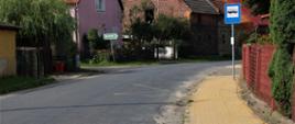 Asfaltowa droga oraz chodnik i zabudowania we wsi. 