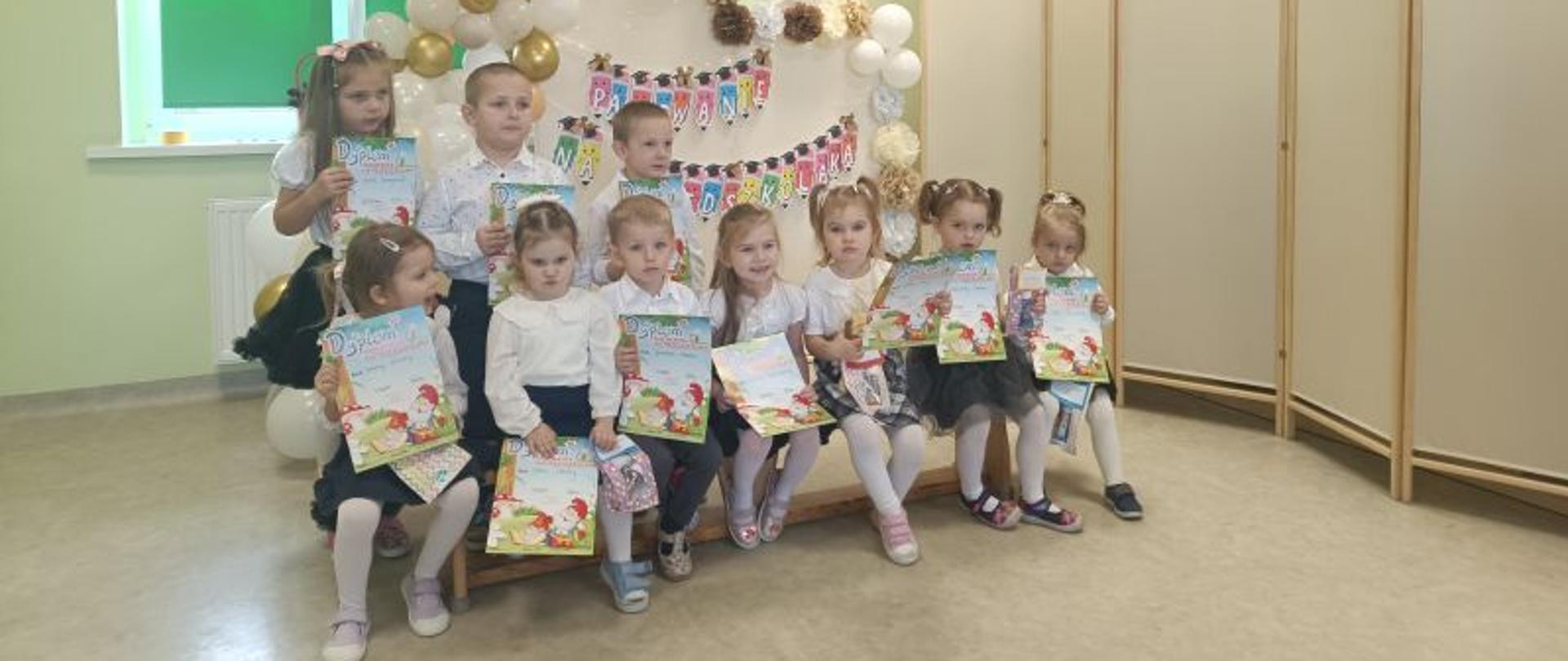 Grupa przedszkolaków, siedem dzieci siedzi, troje stoi, w rękach trzymają dyplomy. Za nimi ścianka dekoracyjna a na niej napis Pasowanie na Przedszkolaka. 
