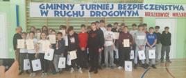 Liczna grupa dzieci i młodzieży pozująca do wspólnej fotografii po zakończeniu "Gminnego Turnieju Bezpieczeństwa w Ruchu Drogowym" w Nieszkowicach Wielkich. Wielu uczestników z dumą prezentuje otrzymane dyplomy, puchary oraz białe torby z upominkami, stojąc na tle pamiątkowego transparentu w szkolnej hali sportowej.