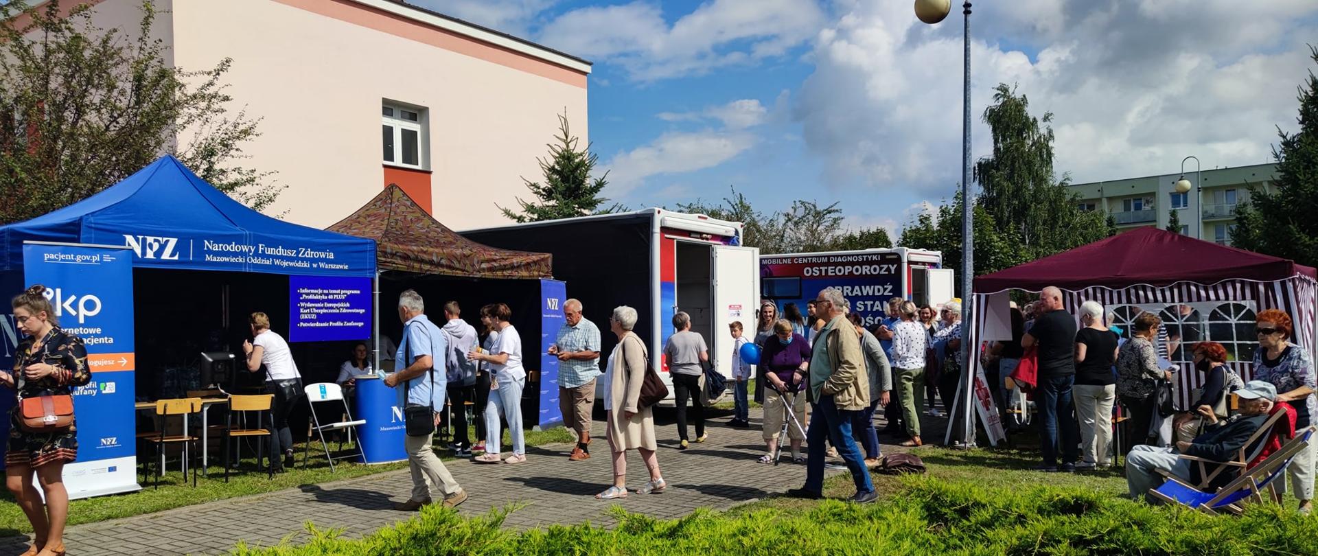 Zdjęcie przedstawia namioty wystawiennicze Narodowego Funduszu Zdrowia, Caritasu, dermobus, osteobus oraz przybyłych na festyn uczestników. Fotografia wykonana na Festynie Profilaktyczno Zdrowotnym na terenie ogrodów przasnyskiego szpitala.