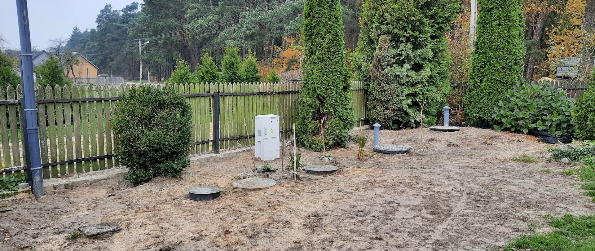 Zdjęcie przedstawia wkopane zbiorniki służące do oczyszczania ścieków. Wokół znajdują się krzewy i płot.