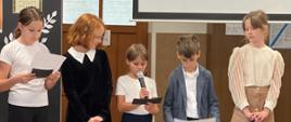 Na scenie znajduje się pięcioro uczniów- cztery dziewczynki oraz chłopiec. Dziewczynka, stojąca w środku przemawia przez mikrofon. 