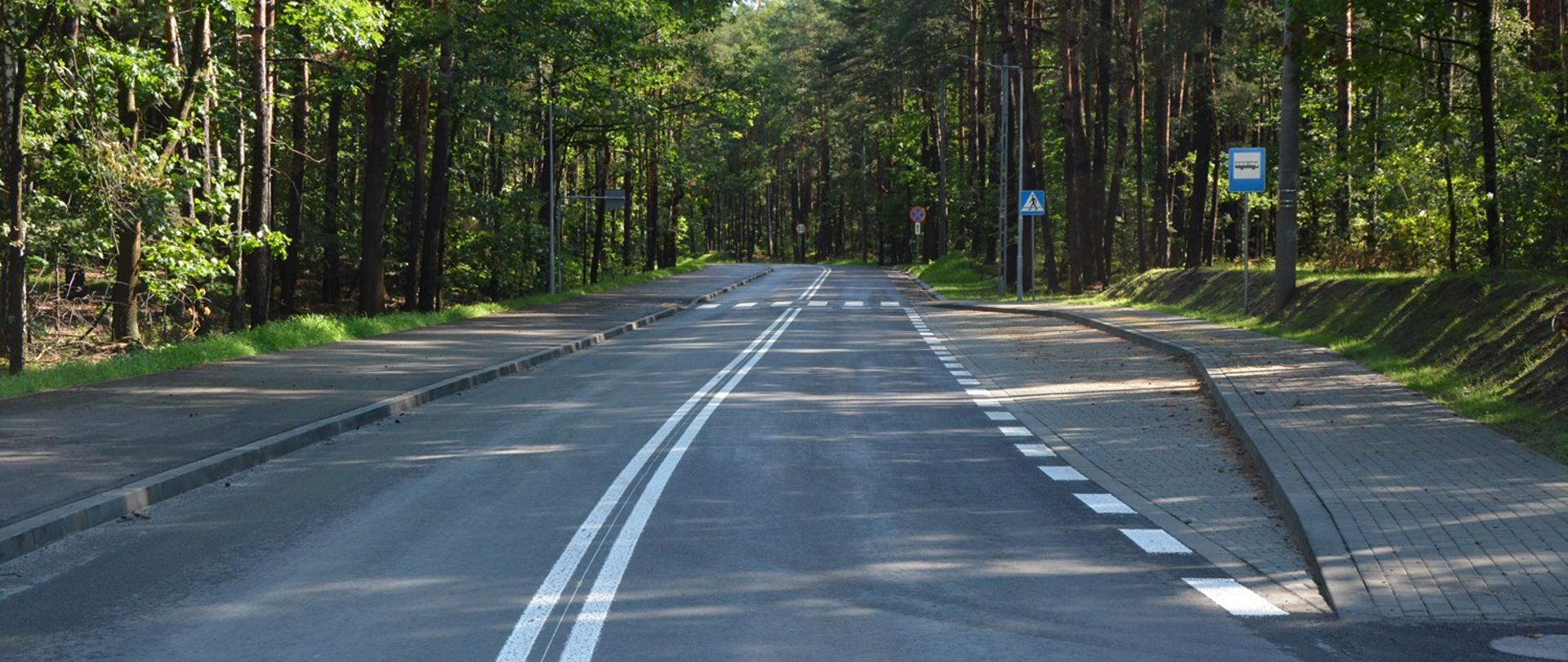 nowa nawierzchnia drogi, na której narysowane są białe linie, z prawej strony zatoczka autobusowa, po obu stronach las 