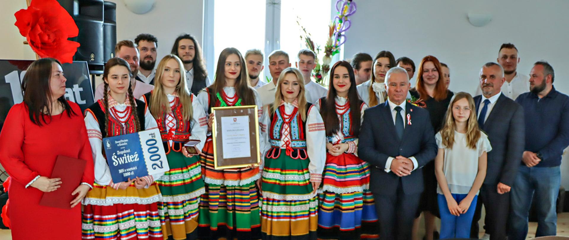 Zdjęcie przedstawia przedstawicieli Zarządu Powiatu w Kraśniku oraz członków zespołu pozujących do pamiątkowego zdjęcia. Członkowie zespołu ubrani są w kolorowe stroje ludowe.