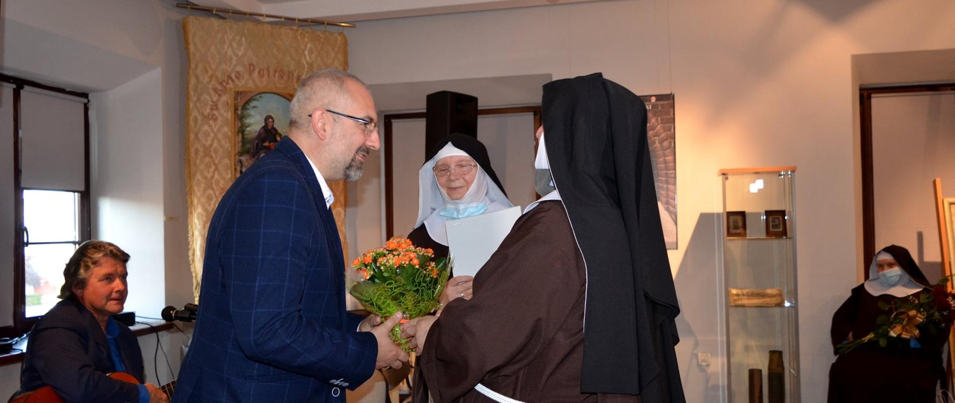 Spotkanie promujące książkę sióstr Honoraty Zofii Szwarc oraz Immakulaty Marii Klickiej „Okres II wojny światowej we wspomnieniach Sióstr Klarysek Kapucynek z Przasnysza". Zdjęcie wykonano w galerii Muzeum Historycznego w Przasnyszu. Fotografia przedstawia Starostę Przasnyskiego Krzysztofa Bieńkowskiego wręczającego bukiet kwiatów Siostrom Klaryskom: Honoracie Zofii Szwarc i Marii Klickiej.