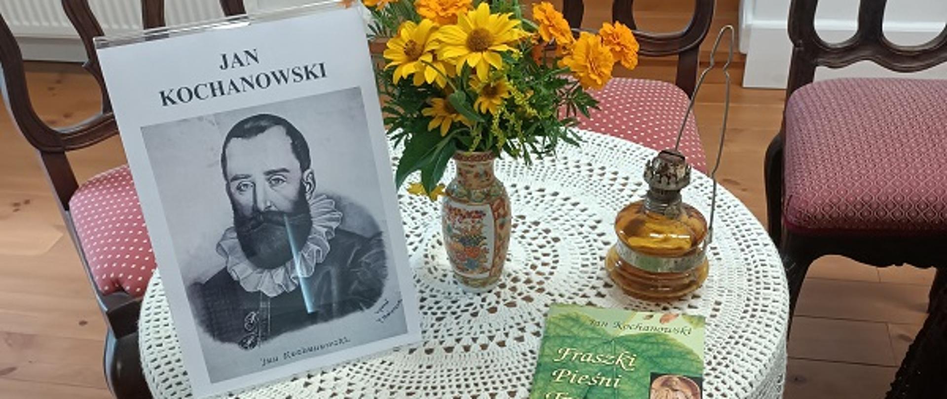 okrągły stół nakryty białą serwetą. na nim tomik poezji, fotografia Jana Kochanowskiego, bukiet polnych kwiatów i lampa naftowa