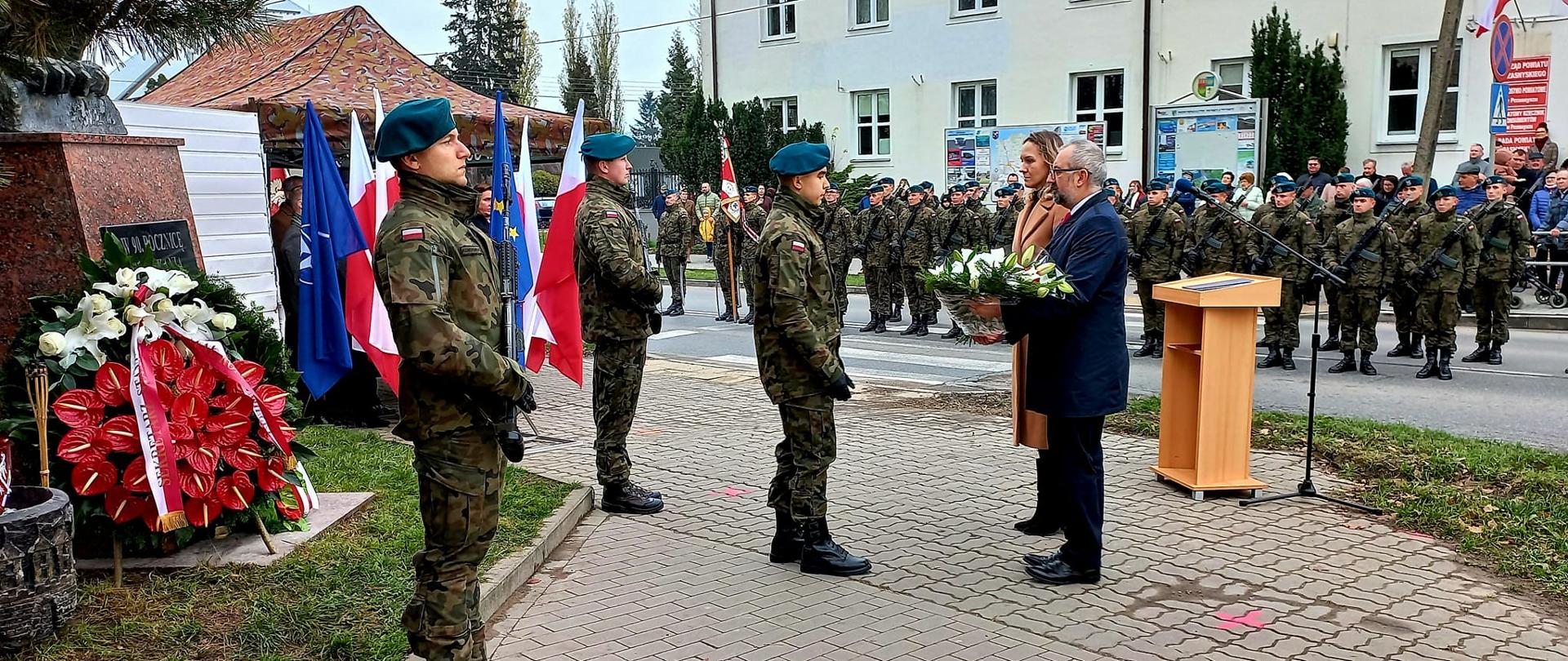 Zdjęcie przedstawia dwóch żołnierzy pełniących wartę pod pomnikiem Niepodległości Polski, podczas gdy trzeci odbiera od Starosty Przasnyskiego Krzysztofa Bieńkowskiego i Przewodniczącej Rady Powiatu Przasnyskiego Katarzyny Wróbel wieniec kwiatów. W tle mównica, dwa szeregi żołnierzy stojących na baczność, poczet sztandarowy 2. Ośrodka Radioelektronicznego w Przasnyszu oraz zgromadzeni na uroczystości goście i Przasnyszanie.