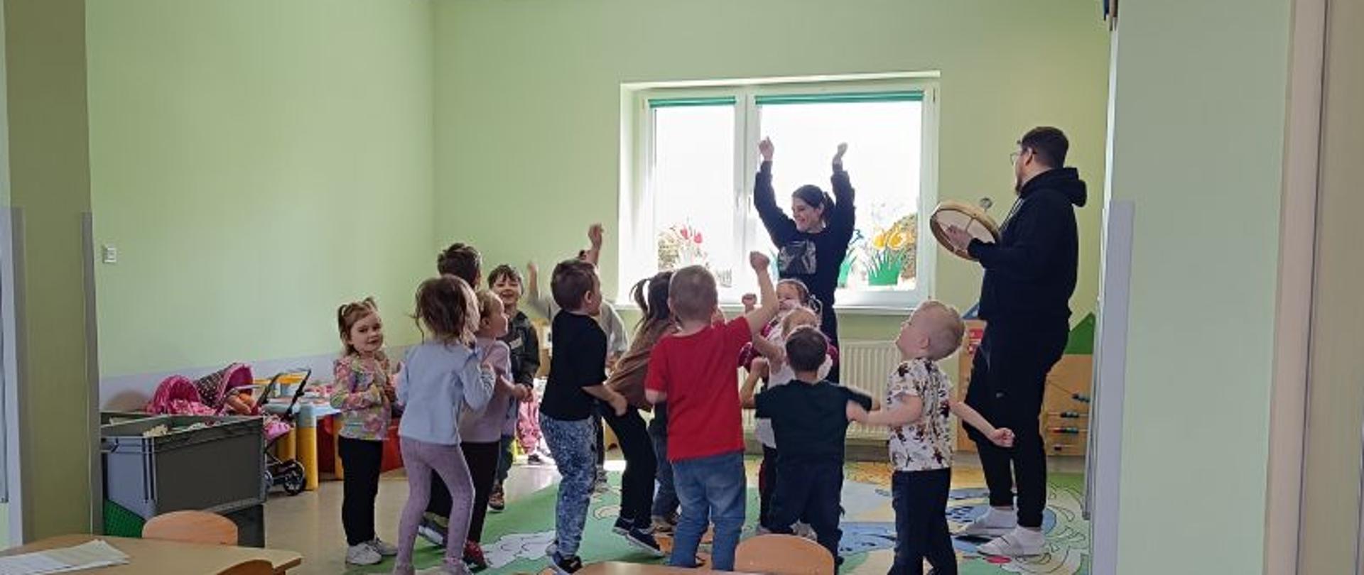 Dzieci w wieku przedszkolnym większość widoczna z tyłu, zwrócone są w stronę kobiety z podniesionymi rękami, obok stoi mężczyzna w czarnym dresie, w rękach trzyma bębenek. Dzieci podskakują, mają podniesioną jedną lub dwie ręce, są radosne. Sala ma ściany w kolorze zielonym, na podłodze znajduje się kolorowy dywan, po prawej stronie znajdują się klocki w skrzyniach, wózki z lalkami. Na wprost widać okno. 