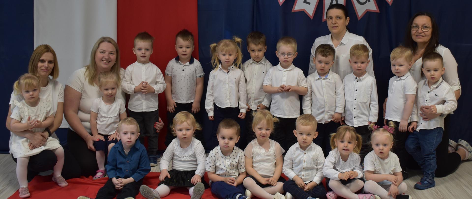 Na zdjęciu są dzieci i panie z grupy Tygryski. Wszyscy ubrani w galowe stroje. W tle widnieje dekoracje o tematyce patriotycznej.