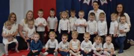 Na zdjęciu są dzieci i panie z grupy Tygryski. Wszyscy ubrani w galowe stroje. W tle widnieje dekoracje o tematyce patriotycznej.