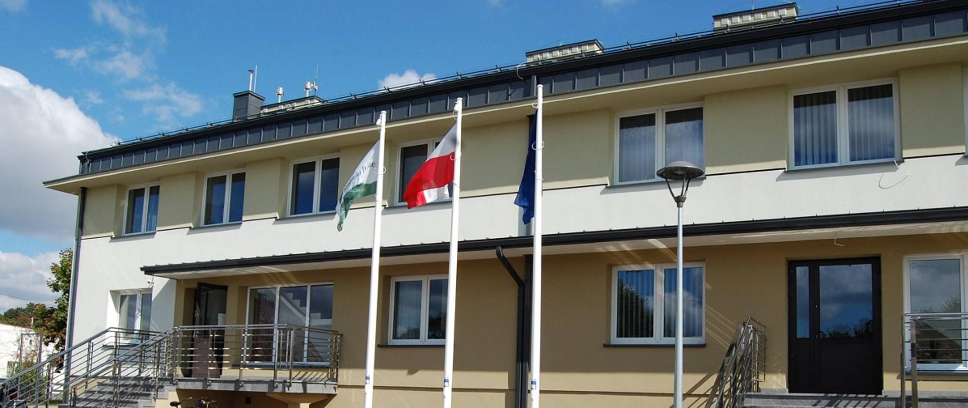 Biało - beżowy budynek z ciemnym dachem. Dwa wejścia po schodach do budynku. Przed budynkiem ławki, stojak na rowery oraz trzy maszty z zielona flagą Gminy Grodzisko Dolne, biało - czerwoną flagą Polski niebieska flagą Unii Europejskiej. 