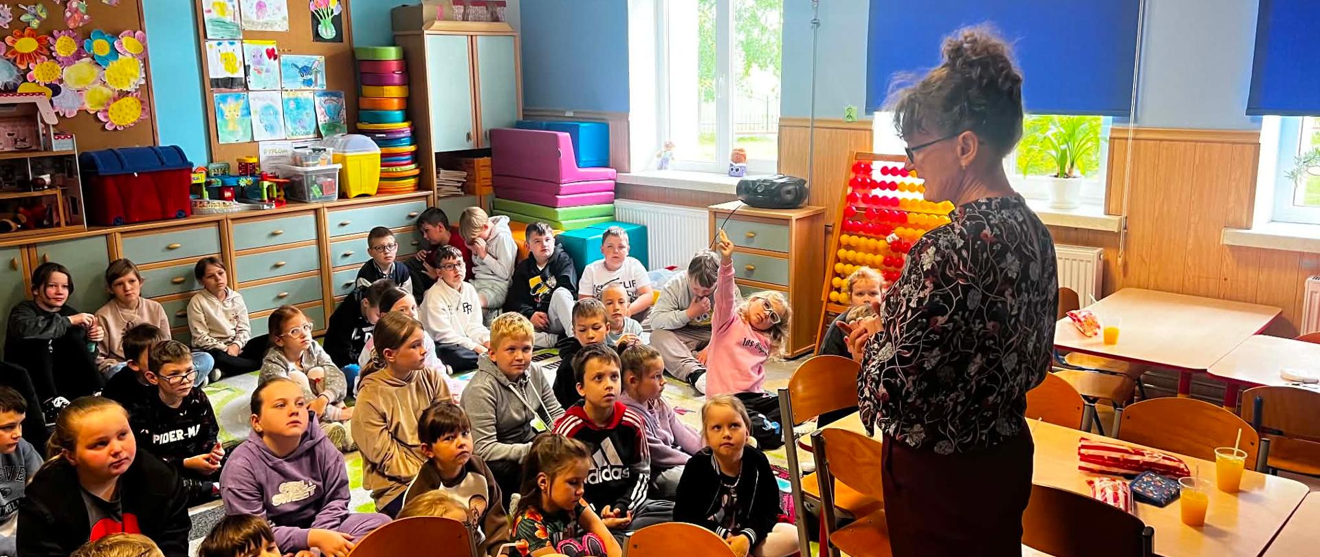 Po prawej stronie kadru stoi tyłem do obiektywu dorosła kobieta w ciemnej bluzce w kwiatowy wzór i ciemnych spodniach, z włosami upiętymi w kok. Kobieta zwraca się do grupy około trzydziestu uczniów w wieku wczesnoszkolnym. Dzieci siedzą na podłodze w kilku rzędach, uważnie patrząc w stronę prowadzącej; jedna dziewczynka w różowej bluzie podnosi rękę do góry. W tle widoczne są kolorowe meble, szafki z pomocami dydaktycznymi, tablice z pracami plastycznymi oraz okna z niebieskimi roletami. 