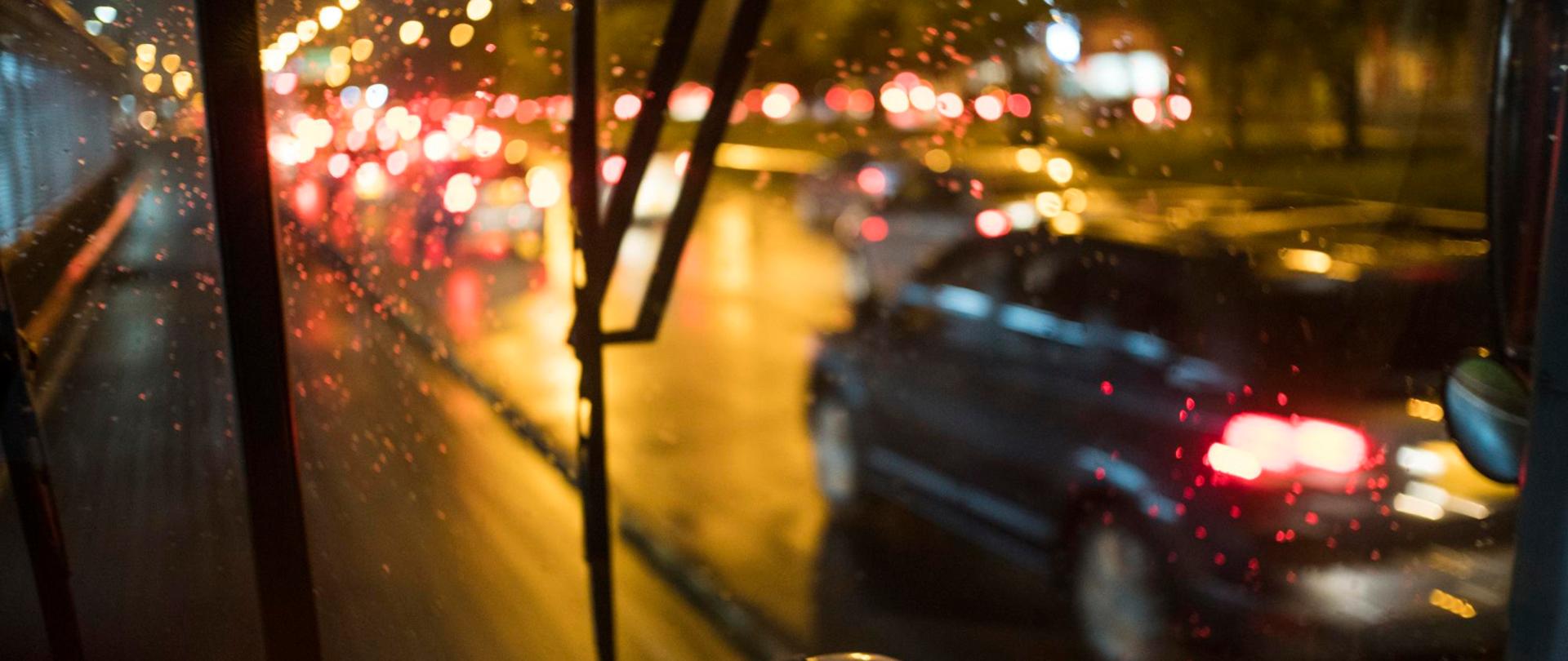 Widok z wnętrza autobusu na nocny ruch uliczny podczas deszczu. Krople wody na szybie, światła samochodów i latarni odbijają się na mokrej jezdni.