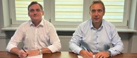 Dwóch mężczyzn w jasnych koszulach siedzi przy drewnianym stole w biurze i podpisuje dokumenty. W tle znajdują się trzy duże okna z roletami dzień–noc. N