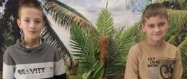 Dwóch uczniów stoi na tle fototapety z palmami, pomiędzy nimi znajduje się mały okrągły stolik, na którym siedzi szynszyla