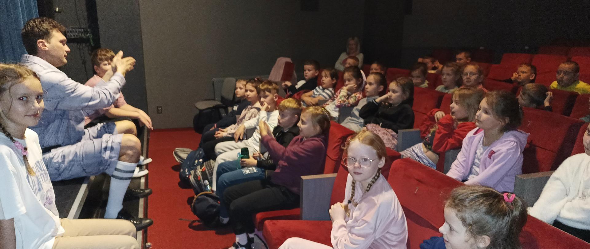 Dzieci siedzące na widowni na czerwonych fotelach. Przed nimi na końcu sceny siedzi mężczyzna unoszący prawą rękę przed siebie i dwie dziewczyny.