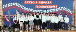 Uczniowie klasy piątej ubrani na galowo, dziewczynki z czerwonymi kokardkami we włosach, chłopcy z czerwonymi kokardkami pod szyją, występują w akademii, za nimi granatowa dekoracja z napisem "Cześć i chwała bohaterom" oraz biało-czerwonymi listkami i wizerunkiem wilka na środku, po lewej stronie cztery brzozowe krzyże ozdobione biało czerwonymi flagami, przy nich wykonane z szarego papieru kamienie, kwiaty i znicze