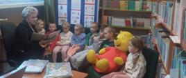 Bibliotekarka czyta przedszkolakom książkę o Kubusiu Puchatku, dzieci siedzą na kanapie w bibliotece, między nimi duży żółty miś w czerwonym sweterku, z tyłu regały z książkami
