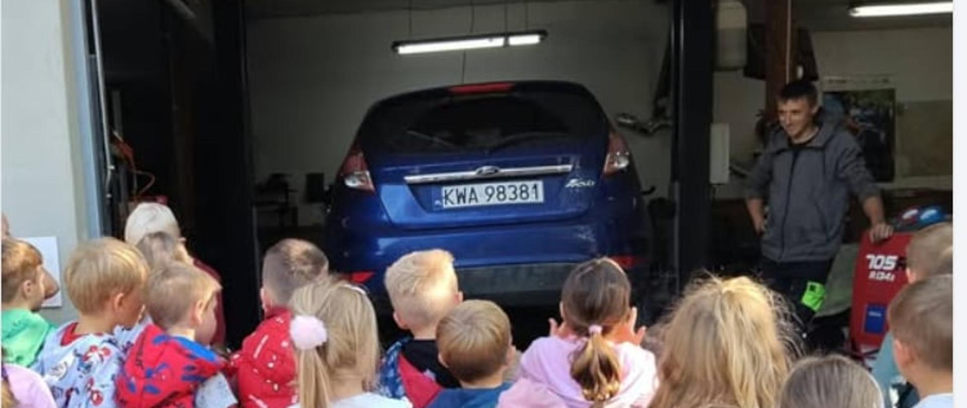 Dzieci z zaciekawieniem przyglądają się samochodowi uniesionemu na podnośniku.