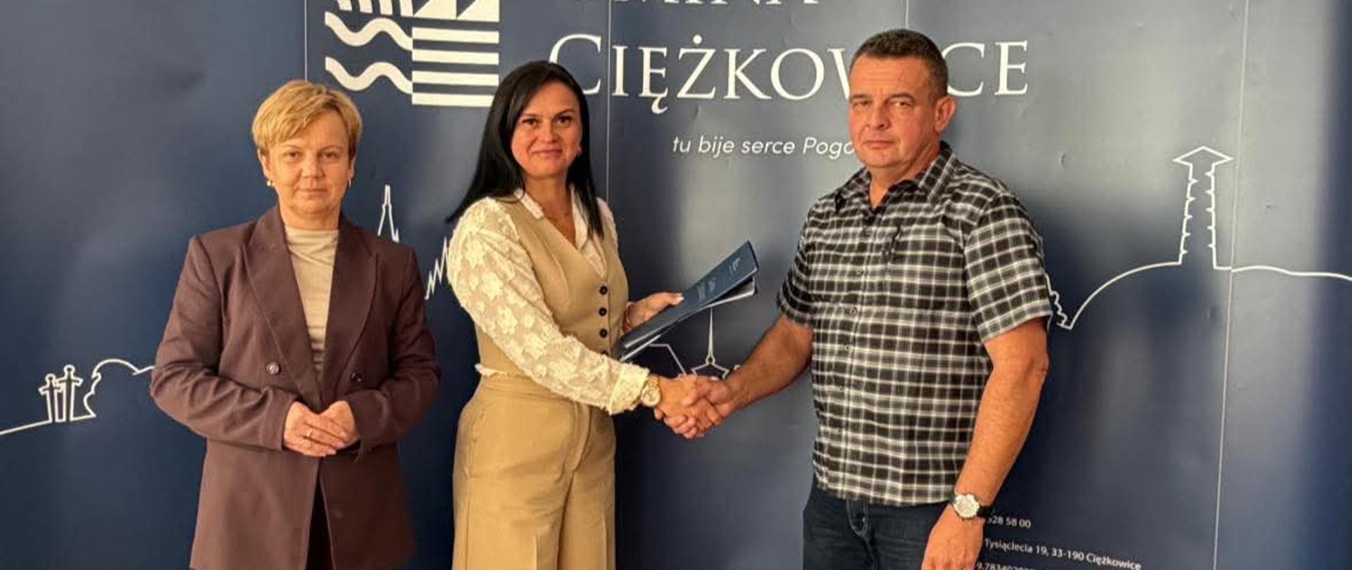 Trzy osoby stojące na tle granatowej ściany z napisem gmina Ciężkowice