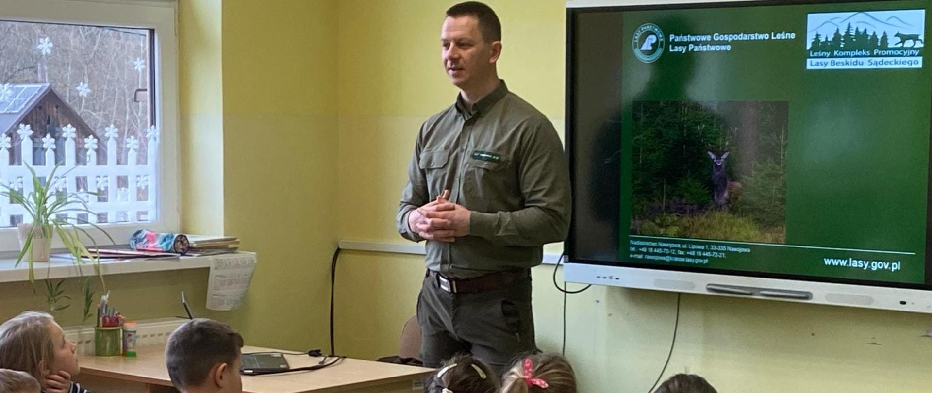 Dzieci siedzą przy ławkach szkolnych, zwrócone są twarzą do tablicy multimedialnej, na której leśniczy pan Przemysław Nosal wyświetla prezentację na temat lasów państwowych