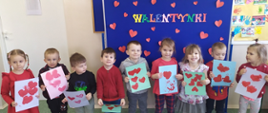 Zdjęcie przedstawia dziewięcioro dzieci ustawionych w szeregu z grupy trzy-latków. Dzieci trzymają w rękach prace na kartkach z serduszkami dla rodziców z okazji walentynek. Za dziećmi niebieska tablica z napisem walentynki. Dookoła napisu przyklejona małe czerwone serduszka. 