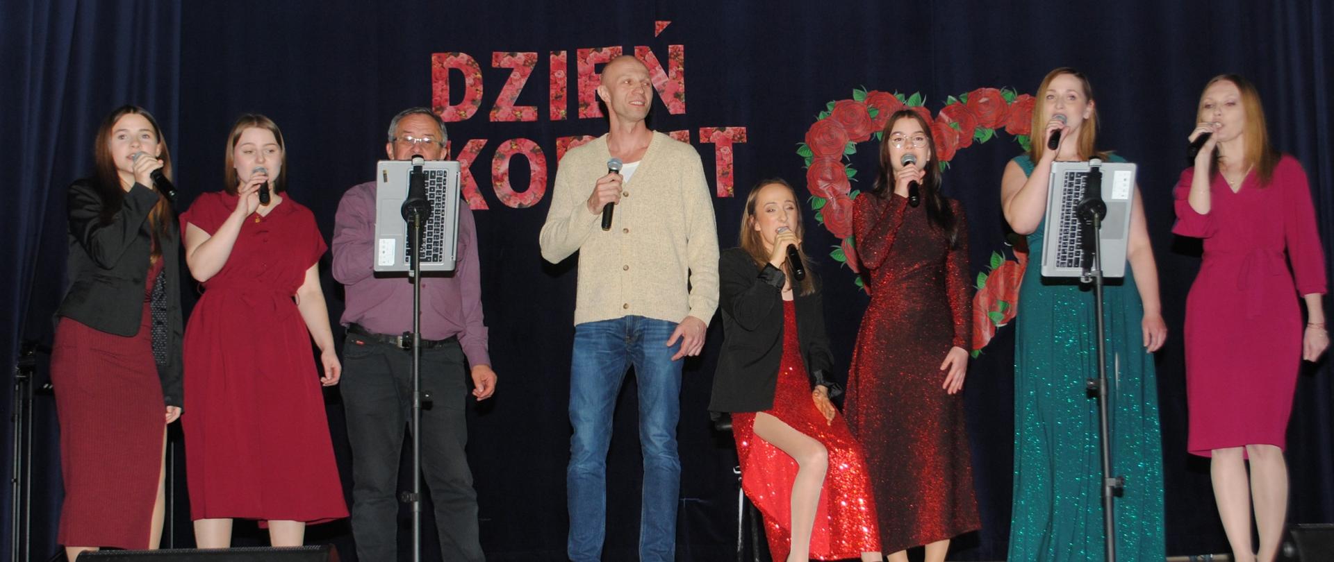 Zdjęcie przedstawia 6 pań i dwóch panów śpiewających podczas występu na dzień kobiet w sali widowiskowej w Ośrodku Kultury.
