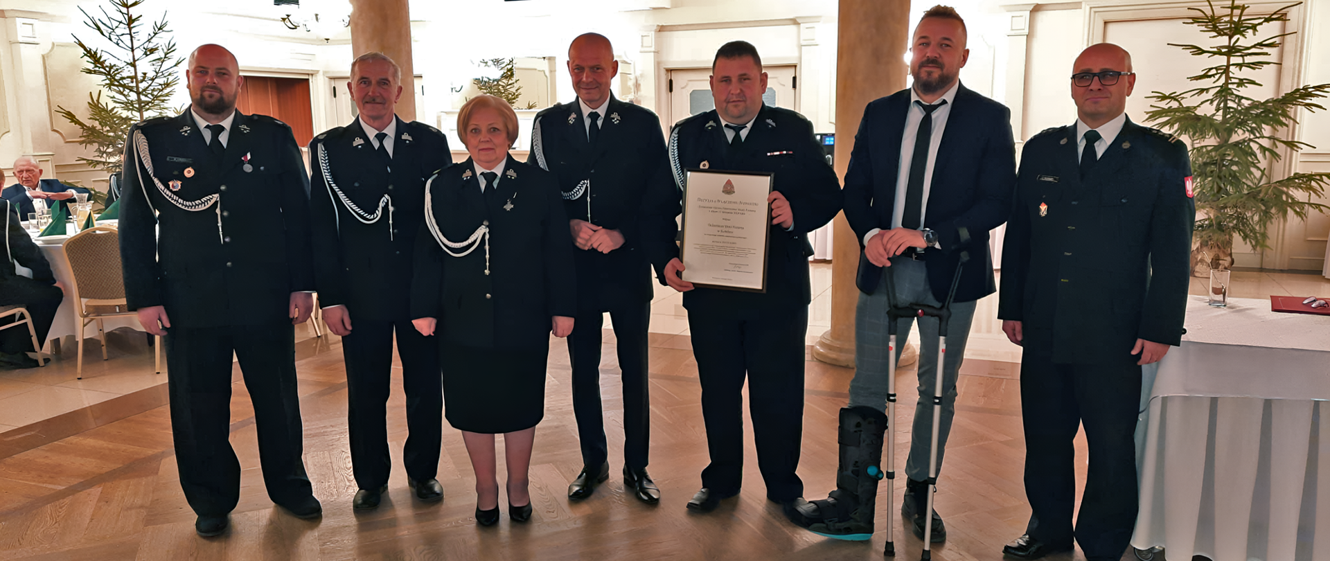 Osoby w mundurach strażackich pozują do zdjęcia w jasnej sali bankietowej.