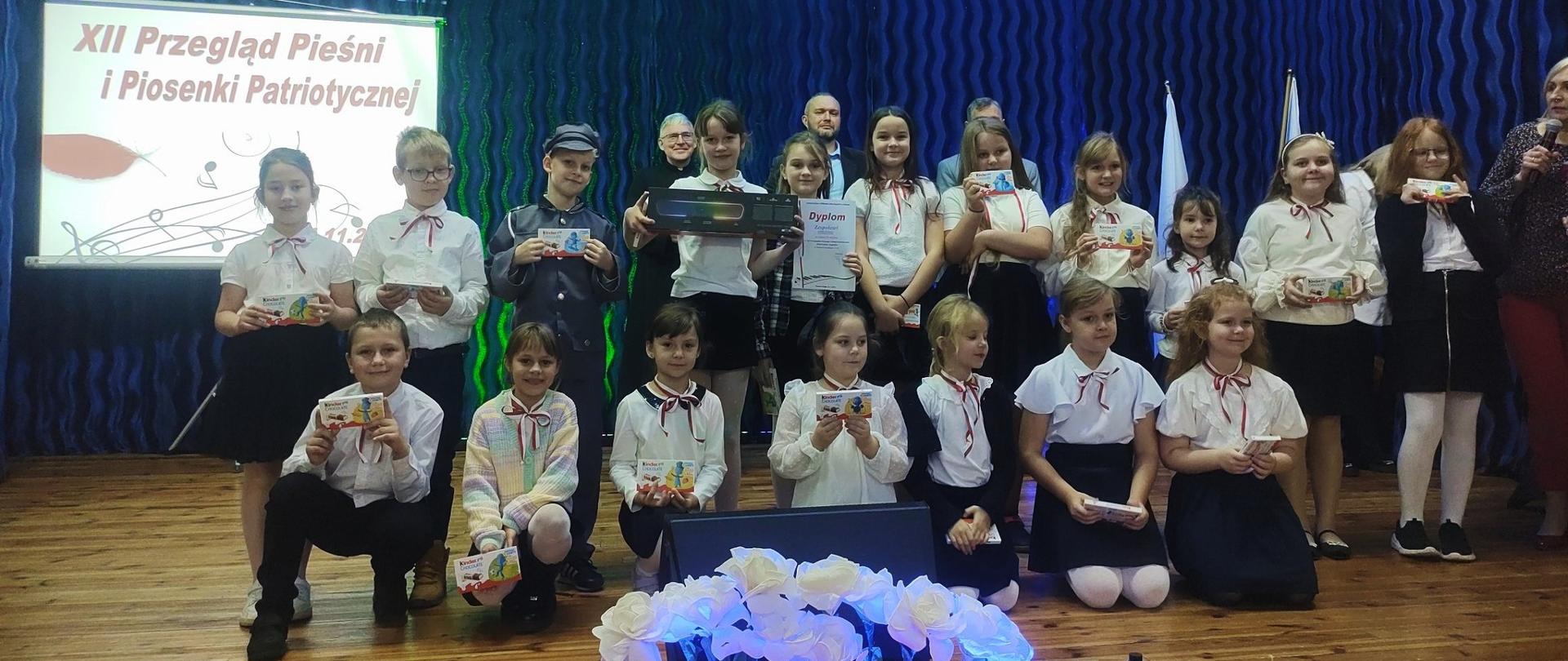 Na scenie znajduje się kilkanaścioro uczniów klas młodszych. Wszyscy ubrani są na galowo. Każdy ma w dłoni czekoladę. Jedna z dziewczynek trzyma w dłoniach głośnik, a druga, stojąca obok niej - dyplom.