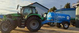 Zielony ciągnik Deutz-Fahr ciągnie niebieski zbiornik na wodę pitną o pojemności 5000 litrów, ustawiony na placu obok budynków gospodarczych.