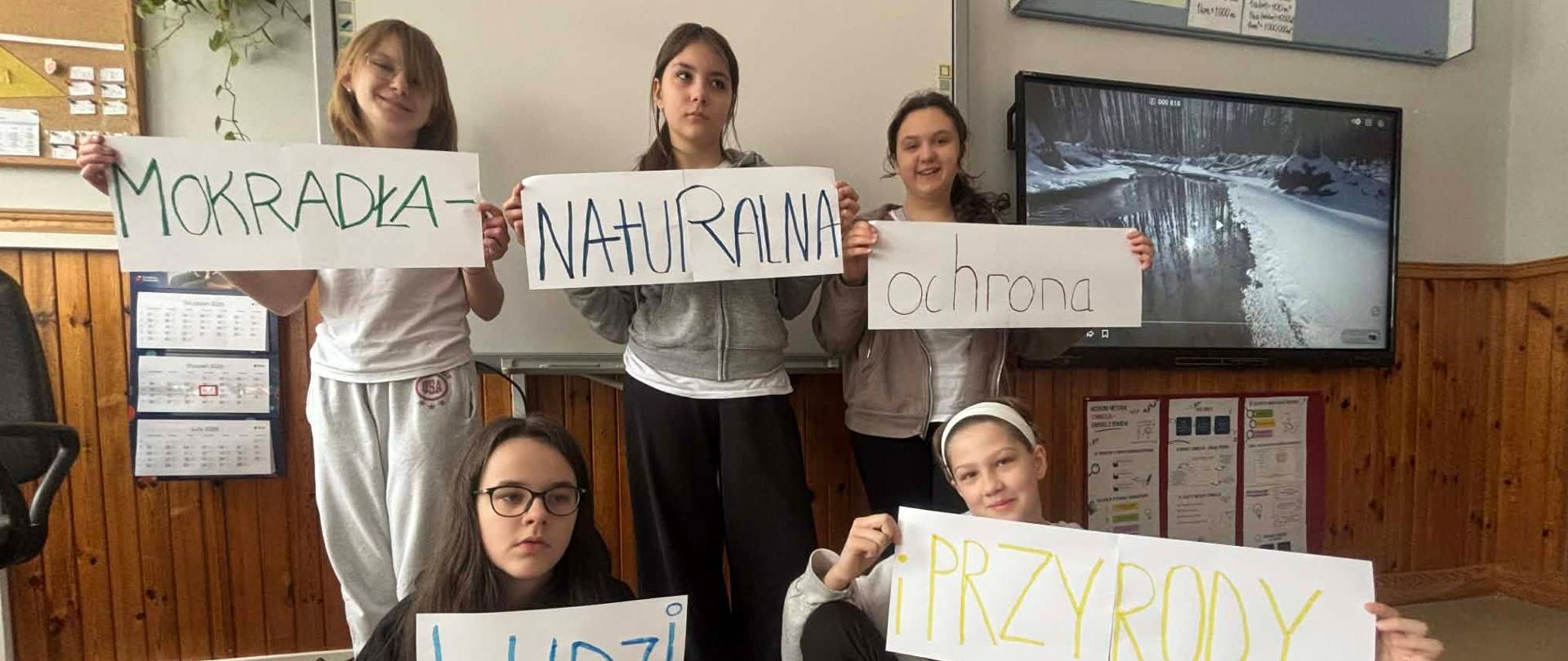 Pięć uczennic klasy szóstej pozuje do zdjęcia z kartkami, które wspólnie tworzą napis: ,,MOKRADŁA - NATURALNA OCHRONA LUDZI I PRZYRODY''.