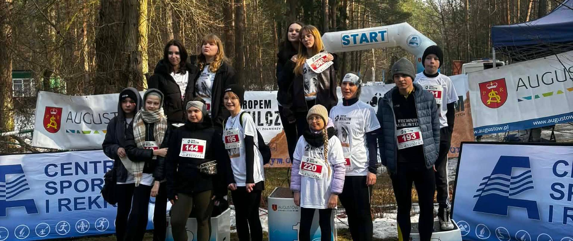 Na zdjęciu znajduje się kilkunastu uczniów naszej szkoły biorących udział w bieg upamiętniającym Żołnierzy Wyklętych. Uczniowie mają na sobie białe koszulki oraz numerki. Wszyscy pozują do wspólnego zdjęcia.