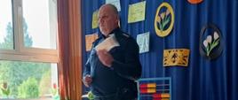 Przy stole nakrytym szafirowym płótnem stoi przedstawiciel Policji, który prezentuje rodzicom akcesoria służące rozprowadzaniu i przechowywaniu środków psychoaktywnych