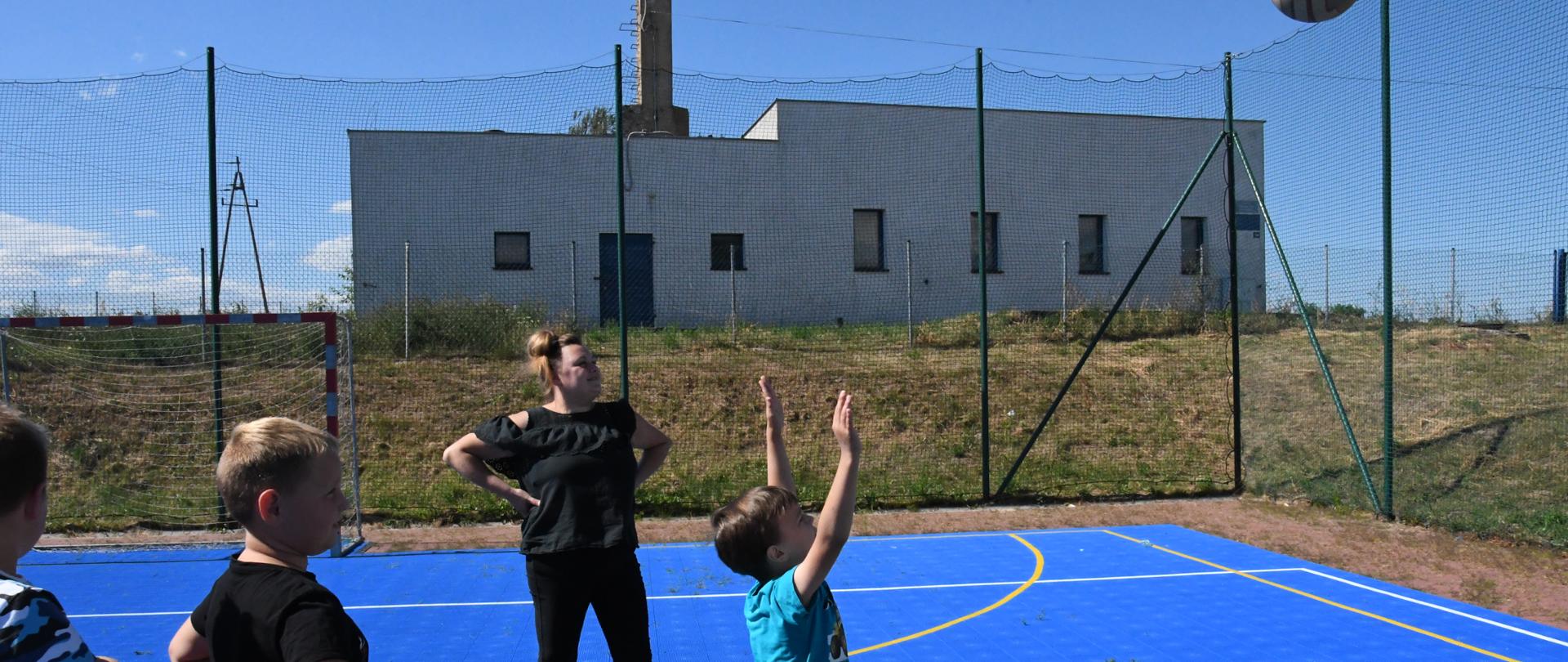 Troje chłopców i jedna kobieta stoją na boisku do piłki nożnej. Chłopiec rzuca do kosza.