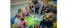 Zdjęcie przedstawia 9 dzieci z nauczycielem prowadzącym Zajęcia z robotyki. Dzieci siedzą na kolorowych pufach w kole. Na środku rozłożona mata zielona i robot do kodowania Photon oraz tablet. Dzieci patrzą na urządzenia a nauczyciel pokazuje kodowanie.