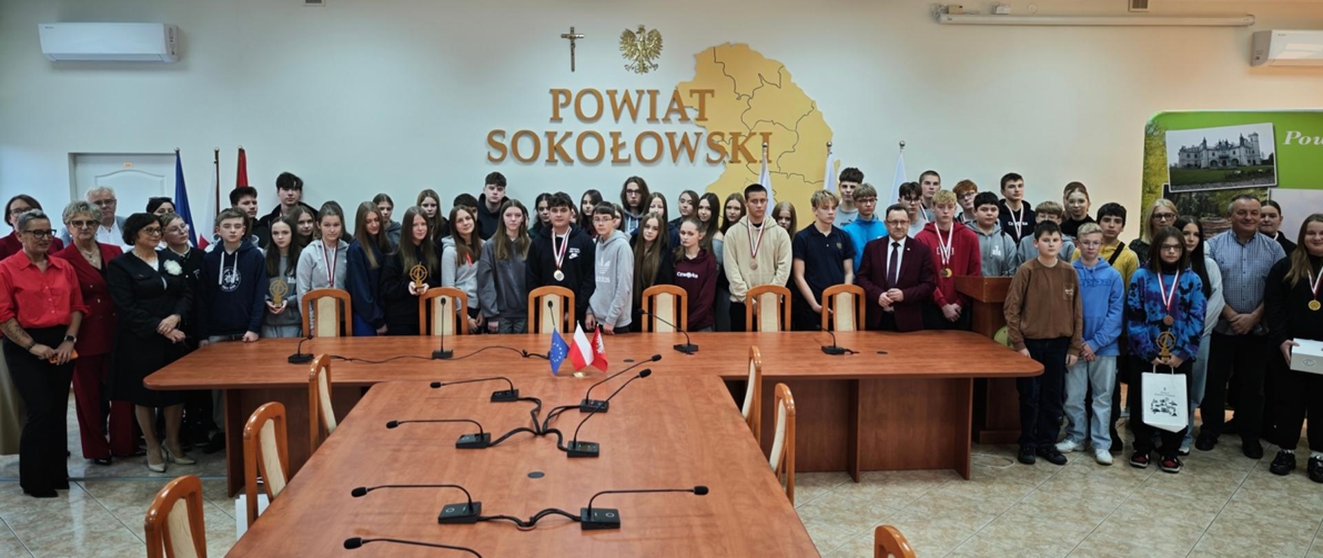 Zdjęcie wykonane w sali konferencyjnej Starostwa Powiatowego w Sokołowie Podlaskim uczestników Młodzieżowych Zawodów Strzeleckich z okazji Święta Niepodległości 2025 o Puchar Starosty Sokołowskiego i Burmistrza Miasta Sokołów Podlaski. Wszyscy stoją na tle napisu Powiat Sokołowski. 