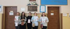 Uczennice trzymają w ręku dyplomy i papierowe torebki z nagrodami, stoją pod ścianą, nad nimi tablica pamiątkowa z nadania imienia szkole, kwiaty i obraz patronki szkoły świętej Kingi
