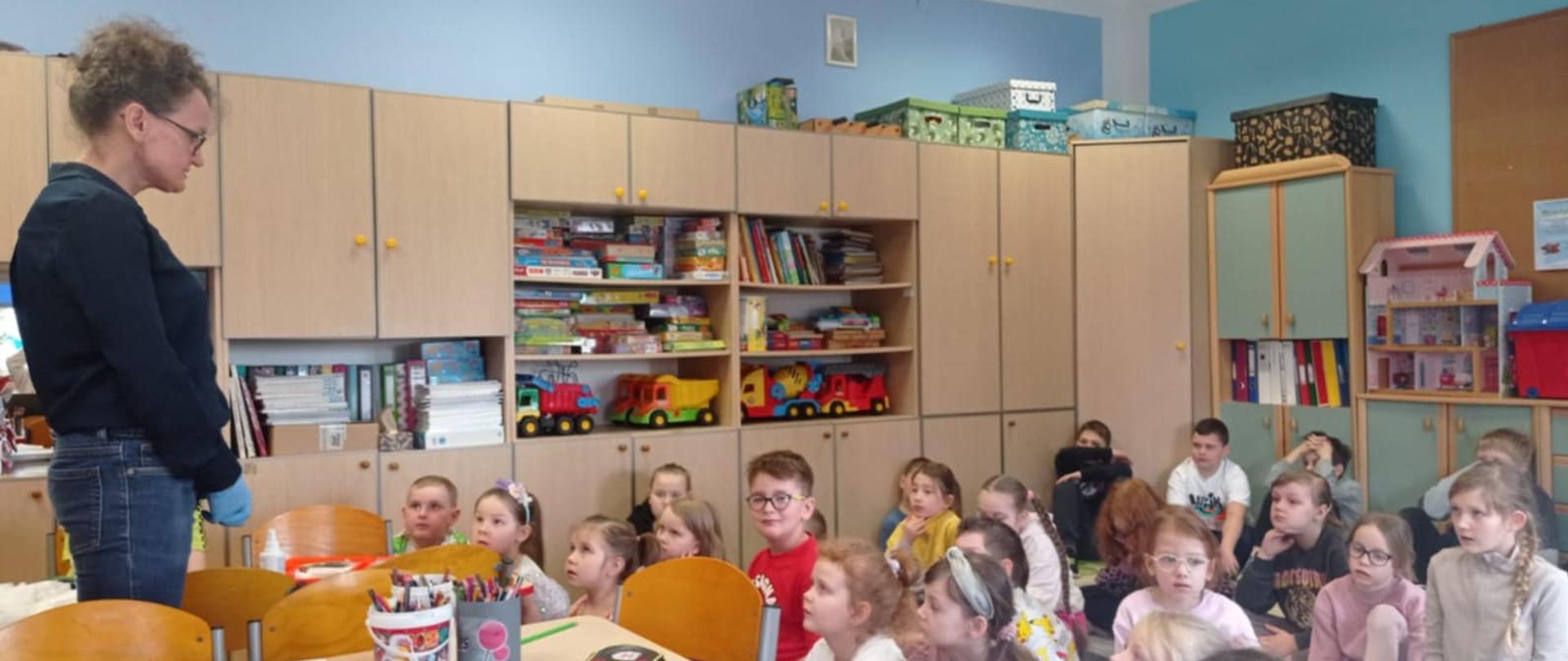 Dzieci z Oddziałów Przedszkolnych oraz klas młodszych siedzą na dywanie. Przed nimi stoi Pani pielęgniarka.