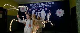 Ubrane w białe bluzeczki i białe spódniczki dziewczynki wykonują taniec wymachując przy tym wstążkami z wbudowanymi małymi światełkami, w tle na granatowej zasłonie wycięte z białego papieru postaci babci i dziadka z wnuczką i wnukiem w ramce utworzonej z różowych serduszek, nad nimi napis "Babciu, dziadku, kochamy Was"