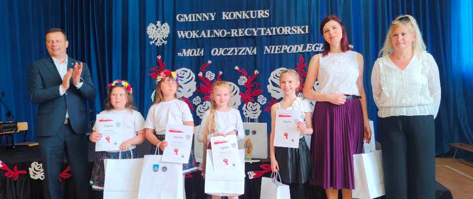 4 dziewczynki w białych bluzkach trzymające papierowe białe torby, obok nich mężczyzna i 2 kobiety