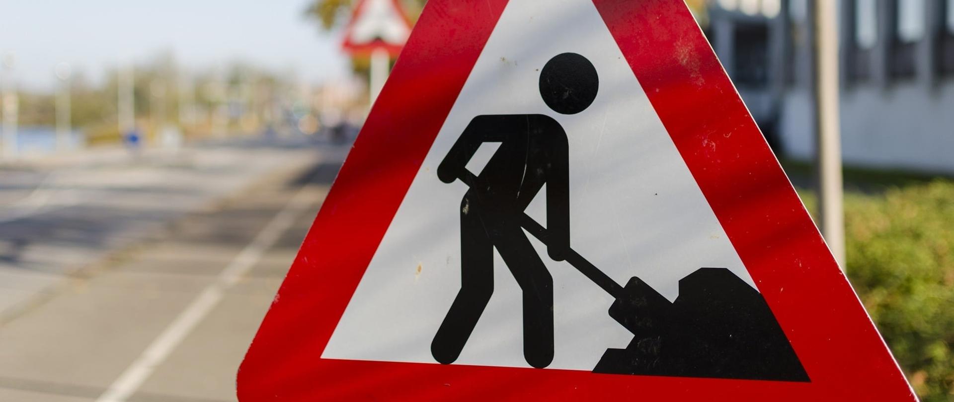 Zdjęcie przedstawia znak drogowy z wyrysowaną na nim postacią, która trzyma w ręce łopatę i wykonuje roboty drogowe. Znak ustawiony na zewnątrz przy drodze.