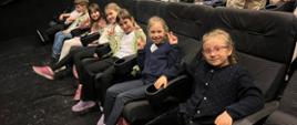 Dzieci siedzą w rzędzie na fotelach widowni teatralnej, uśmiechają się i pokazują znak „V”, czekając na rozpoczęcie spektaklu.